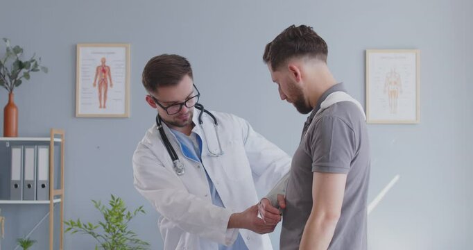 Male young doctor traumatologist examining man patient with broken arm in medical office in exam room. Person with injured hand standing in clinic and having consultation. 4k video.