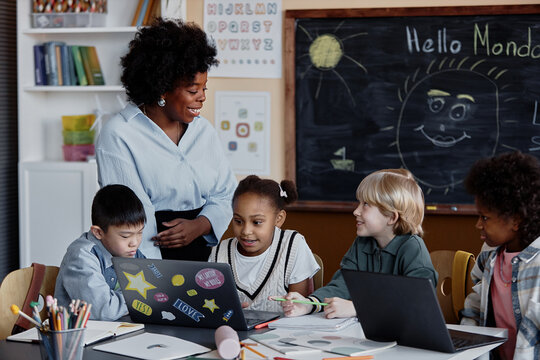 Empathic female teacher smiling answering kids question while conducting lesson in primary school with use of laptops in diverse group of children, copy space