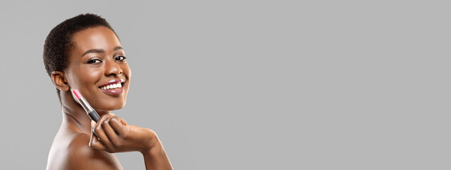 Beauty and cosmetics concept. Portrait of attractive african american woman holding pink lipstick in hand and looking to camera, grey studio background, copy space