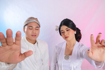 A pair of indonesia men and women with their eyes closed, hands facing forward, wearing modern Javanese traditional clothing, white velvet bridal kebaya, wedding concept with a white studio background