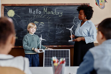 Smiling young boy of primary school showing big solar panel and small wind turbine to classmates answering at blackboard with funny sun drawings