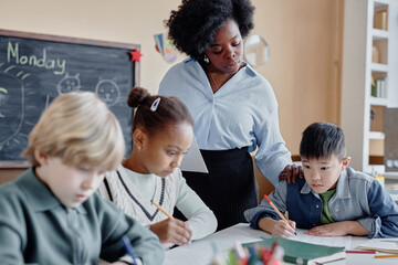 Shot of female teacher of Black ethnicity supervising multiethnic young students writing test or...