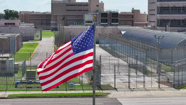 American flag in front of a high-security prison facility. Barbed wire fences, guard towers, and secure buildings. Correctional institution in a city, aerial.