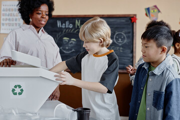 Side view of young boy putting recyclable in waste sorting bin during environmental awareness lesson in multiethnic class