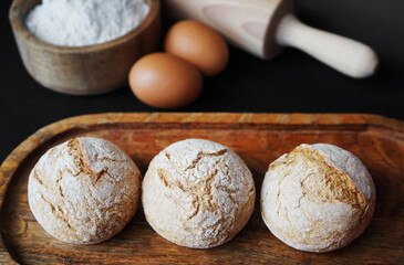 Small fresh breads on a wooden tray next to eggs, a wooden bowl with flour and a rolling pin
