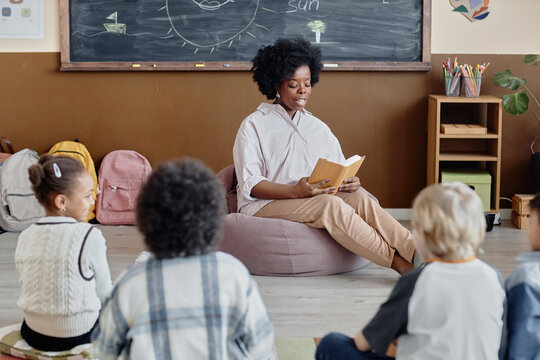 Thoughtful female teacher of Black ethnicity in bean bag chair reading book aloud to primary school students sitting around on classroom floor