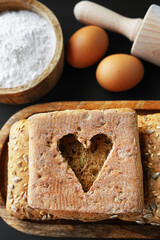 A cut out heart on a slice of bread next to small fresh breads with seeds and sesame on a wooden tray next to eggs, a wooden bowl with flour and a rolling pin