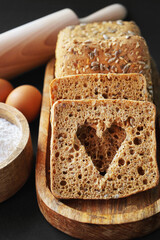 A cut out heart on a slice of bread next to small fresh breads with seeds and sesame on a wooden tray next to eggs, a wooden bowl with flour and a rolling pin