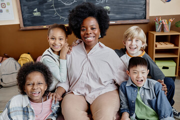 Portrait shot of smiling female teacher of Black ethnicity sitting in chair near multiethnic happy...