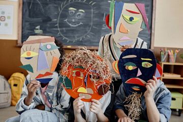 Shot of unrecognizable kids hiding behind cardboard face mask crafts in abstract style in classroom with blackboard