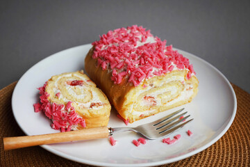 Sponge roll with cream and pink sprinkles on a white plate next to a fork on a gray background