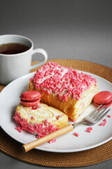 Sponge roll with cream, pink sprinkles and raspberry macaroons on a white plate next to a fork and cup on a gray background