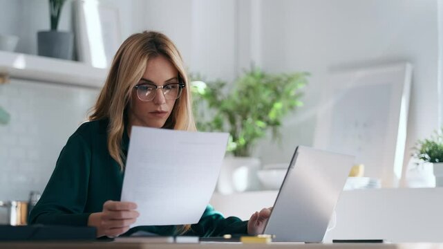 Video of pretty young business woman working with computer while consulting some invoices and documents in the kitchen at home.