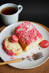 Sponge roll with cream, pink sprinkles and raspberry macaroons on a white plate next to a fork and cup on a dark background