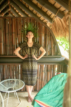 A woman stands confidently under a bamboo shelter, adorned with a unique headdress made of long green leaves, wearing a traditional patterned dress with her hands on her hips.