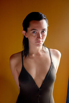 A young woman with wet hair is wearing a dark swimsuit, standing against an orange background, and gazing directly into the camera with a serious expression.