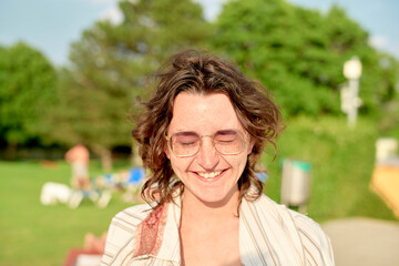 A smiling young woman with curly hair and glasses is standing outdoors on a sunny day, with blurred greenery and people in the background.