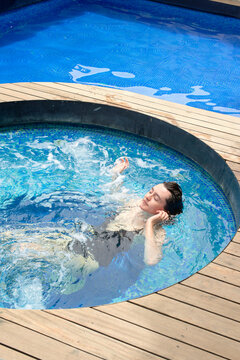 A person is relaxing in a circular whirlpool spa with jets creating bubbles, surrounded by a wooden deck with a larger pool in the background.