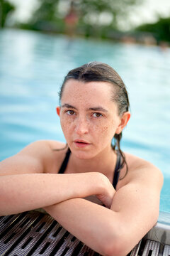 A young woman with wet hair and freckles is resting her arms on the edge of a swimming pool, gazing directly at the camera.