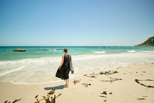 Woman walking alone on a sunny beach with waves in the background and seaweed strewn on the sand.