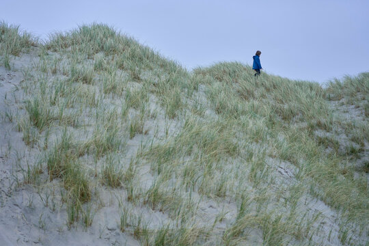 A lone individual sits in contemplation on a grass-covered sand dune under a soft blue sky.