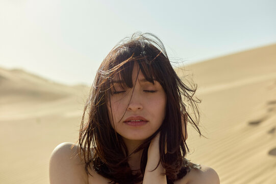 Sun-kissed woman with eyes closed and hair tousled by the wind stands amidst golden desert sands.