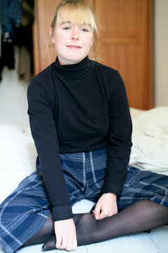 Smiling girl with bangs wearing a black turtleneck and plaid skirt sits on a bed in a cozy room.