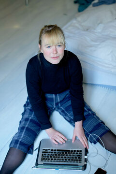 A woman sits on the floor with a laptop on her lap, looking at the camera.