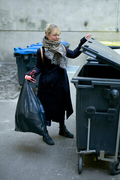 Woman in a blue coat disposing of a trash bag into a grey bin on a city street.