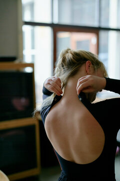 A woman is seen from behind putting on a black sleeveless top, showcasing a refined hairstyle and a bare back against an indoor backdrop.