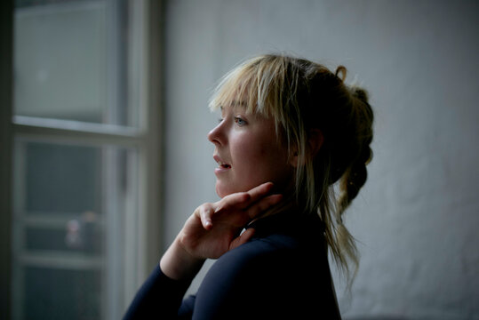 Woman with blonde hair fastening her necklace, standing indoors with natural light coming from a window.