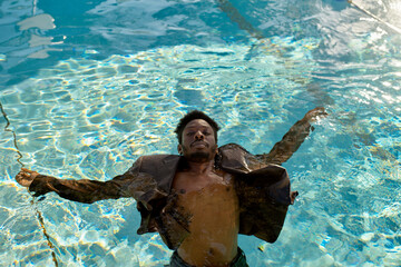 A man calmly floating on his back in a clear swimming pool while wearing a dress shirt.