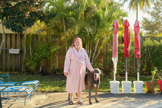 A smiling woman in a bathrobe stands beside a large dog in a sunny backyard with palm trees and folded patio umbrellas in the background.