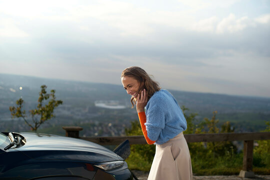 Smiling woman standing by a car overlooks a cityscape from a high vantage point at sunset