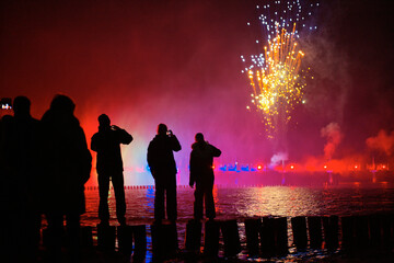 Silhouetted onlookers watch a vibrant fireworks display over a body of water at night.