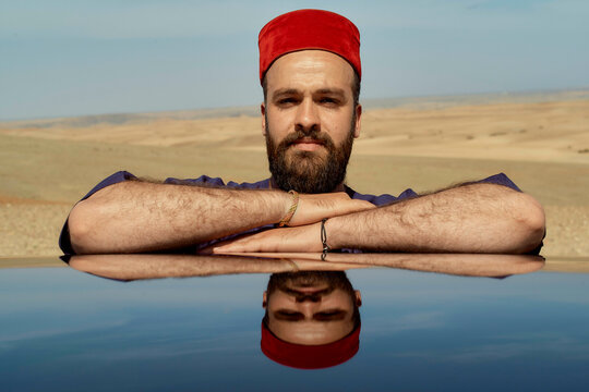 Man with a beard wearing a red fez hat rests his chin on his crossed arms, reflected on a glossy surface against a desert backdrop. - Powered by Adobe