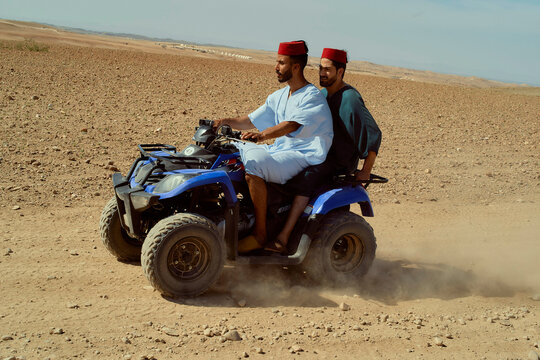 Two men in traditional attire enjoying a quad bike ride through a desert landscape.