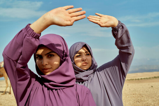 Two women in hijabs posing with their hands framing the sky in a desert landscape.