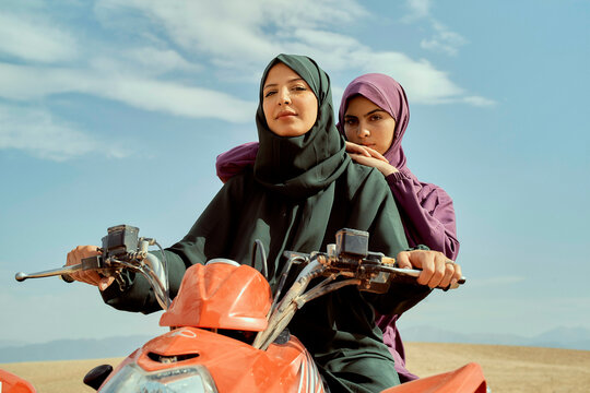 Two women in hijabs riding a red quad bike on sandy terrain under a clear sky.