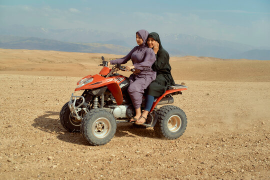 Two young girls smiling while sitting on a red quad bike in a desert landscape with mountains in the background.