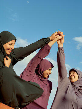 Three women in hijabs holding hands up together in solidarity under a blue sky.