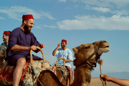 A group of men wearing traditional red hats smiling and riding camels across a desert landscape under a clear sky.