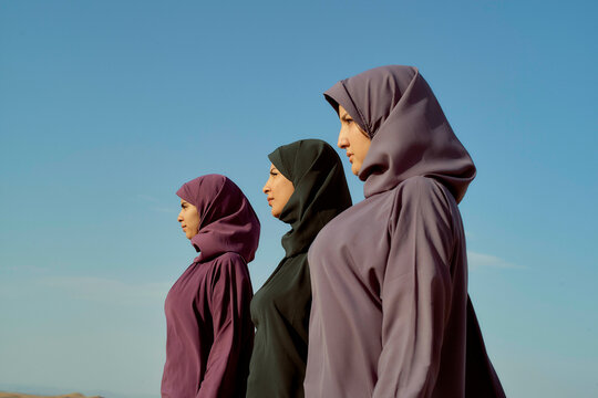 Three women in hijabs standing side by side against a clear blue sky.