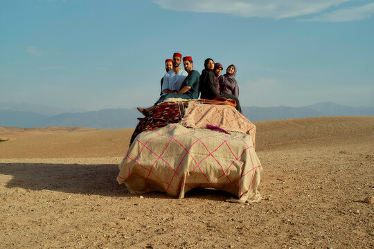 A group of people wearing traditional headgear poses together on an elaborately patterned textile placed on the ground with a backdrop of hilly terrain under a clear sky.
