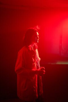 A woman stands in a red-lit room holding a drink, looking contemplatively to the side.