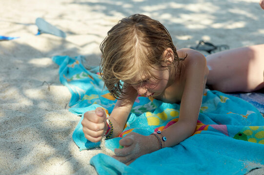 A young child plays contently with sand while lying on a colorful beach towel.