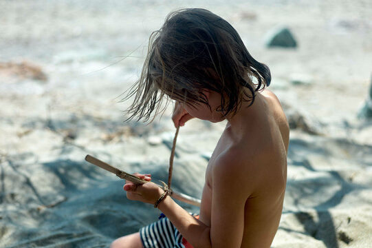 Young child sitting on a sandy beach examining a stick with focused attention under natural sunlight.