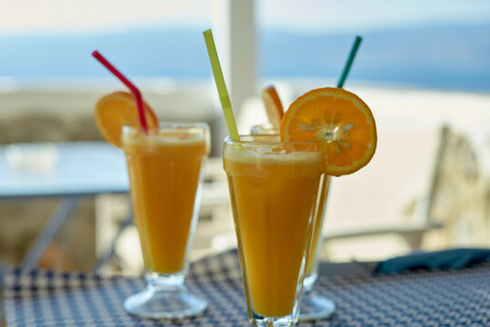 Two glasses of fresh orange juice with straws and an orange slice garnish on a table overlooking a blurred beach view.