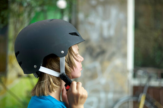 Close-up of a person wearing a black helmet, fastening the chin strap while sitting indoors, with a blurred background.