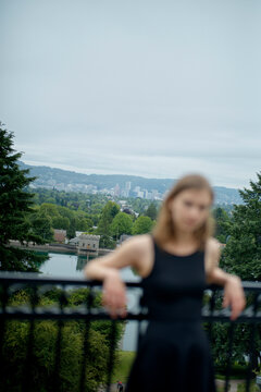 A female in black athletic wear rests on a railing with a blurred city park landscape in the background.